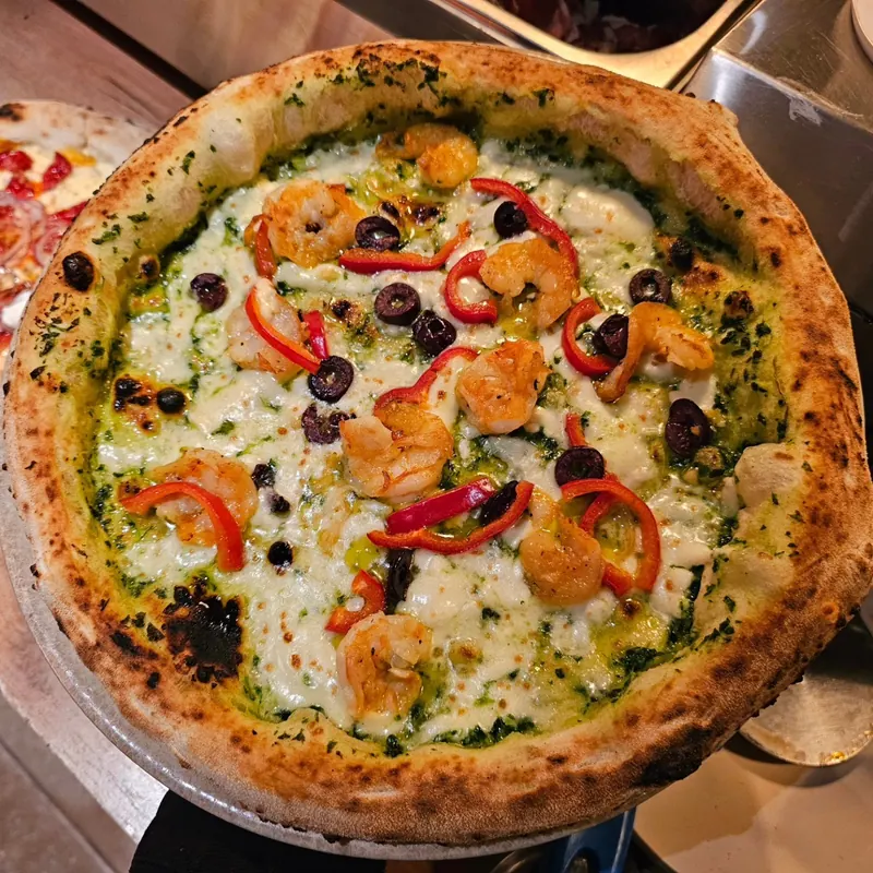 Pizza topped with shrimp, red peppers and black olives on a green pesto base